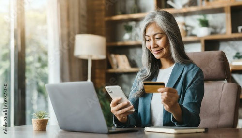 Serene middle-aged grey-haired Asian woman shopping online sitting with credit card and mobile phone at home, multiracial senior 50s female making order online, booking, transfering money  photo