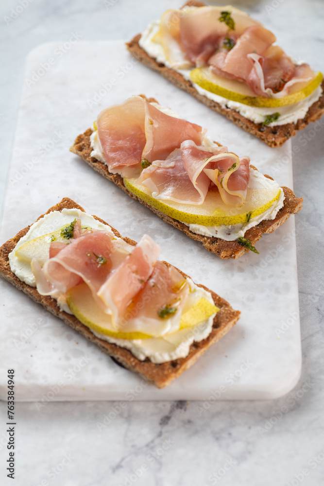 Ideal snack wholewheat crispbread with ricotta cheese, prosciutto, sliced pear and thyme on light grey marble cutting board. Healthy snack concept. Italian cuisine