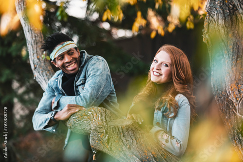Close-up of a mixed race couple smiling and enjoying the outdoors while resting on a tree branch, spending quality time together during a fall family outing in a city park; Edmonton, Alberta, Canada photo