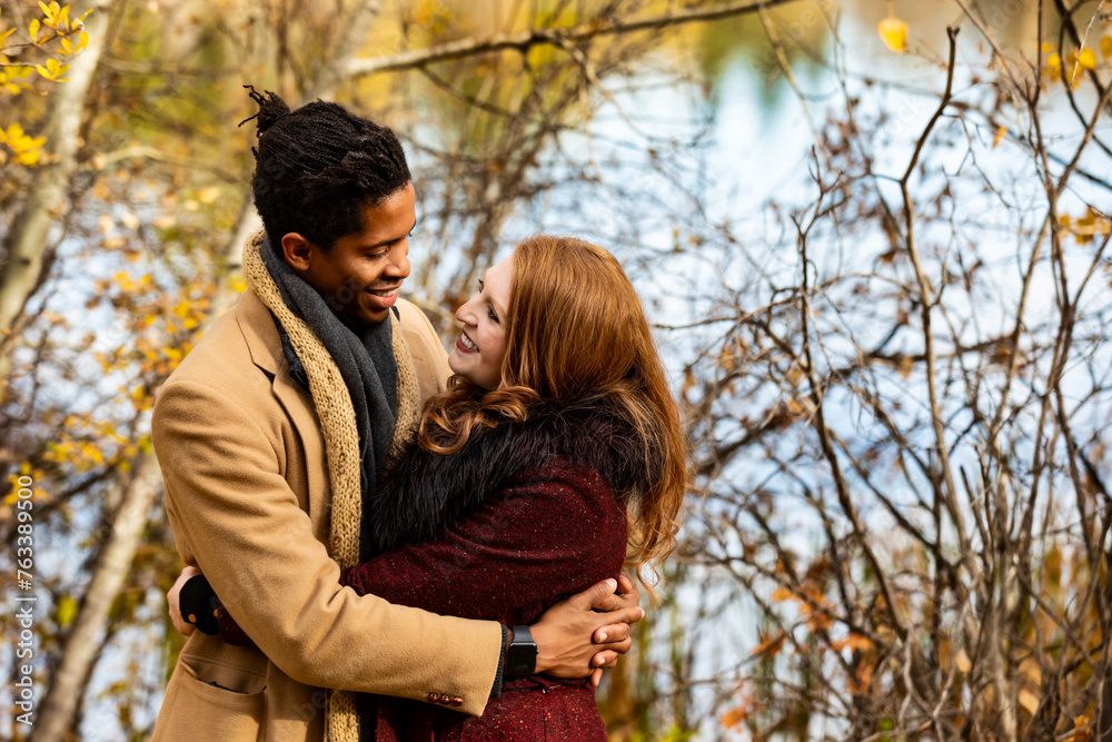 A mixed race couple standing by the shore of a lake hugging each other while spending quality time together during a fall family outing in a city park; Edmonton, Alberta, Canada A mixed race couple standing by the shore of a lake hugging each other while spending quality time together during a fall family outing in a city park; Edmonton, Alberta, Canada