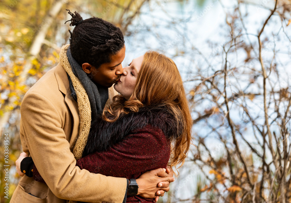 A mixed race couple standing by the shore of a lake hugging and kissing each other while spending quality time together during a fall family outing in a city park; Edmonton, Alberta, Canada A mixed race couple standing by the shore of a lake hugging and kissing each other while spending quality time together during a fall family outing in a city park; Edmonton, Alberta, Canada