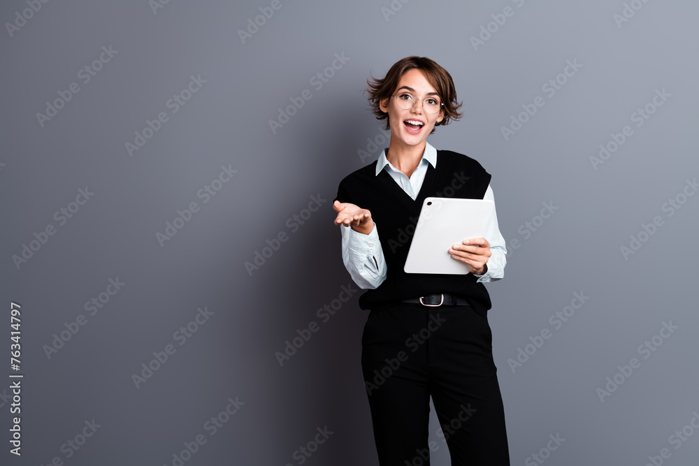 Photo of adorable confident lady wear stylish clothes hold ipad communicate tell report empty space isolated on gray color background Photo of adorable confident lady wear stylish clothes hold ipad communicate tell report empty space isolated on gray color background