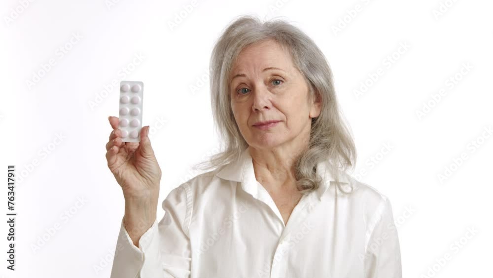 An elderly woman with a contemplative expression holds a blister pack of pills, considering her health options and potential treatment on a white backdrop. Camera 8K RAW. An elderly woman with a contemplative expression holds a blister pack of pills, considering her health options and potential treatment on a white backdrop. Camera 8K RAW.