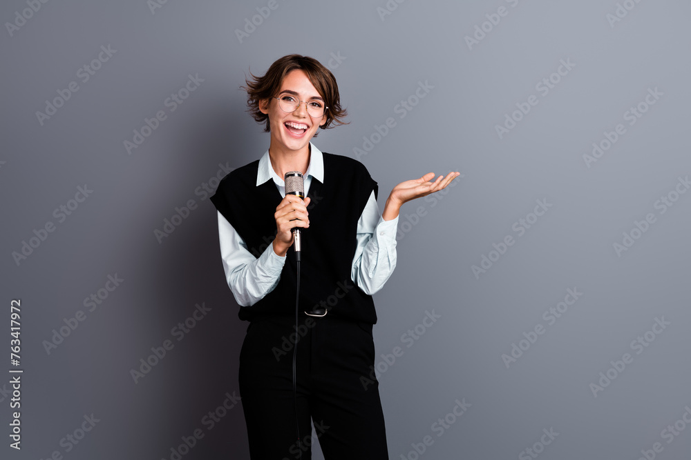 Photo of positive clever woman in glasses talk in microphone presenting profitable offer empty space isolated on gray color background