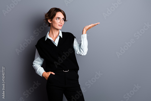 Photo of dreamy cute lady clerk dressed black vest eyewear looking showing arm empty space isolated grey color background © deagreez