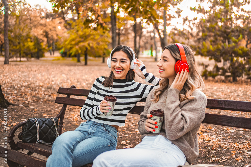 Two women sitting on a park bench with headphones and drinking coffee