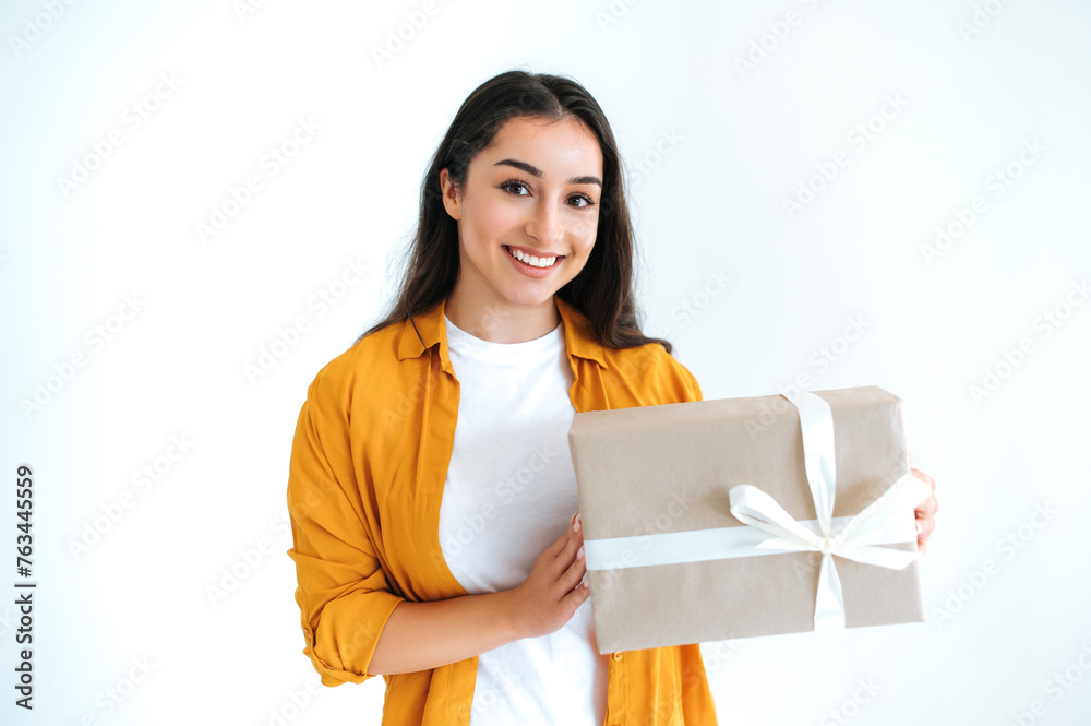Gifts and surprises concept. Excited happy brunette indian or arabian woman in casual orange shirt, stands on isolated white background, holding big gift box, looking at camera and smiles friendly Gifts and surprises concept. Excited happy brunette indian or arabian woman in casual orange shirt, stands on isolated white background, holding big gift box, looking at camera and smiles friendly
