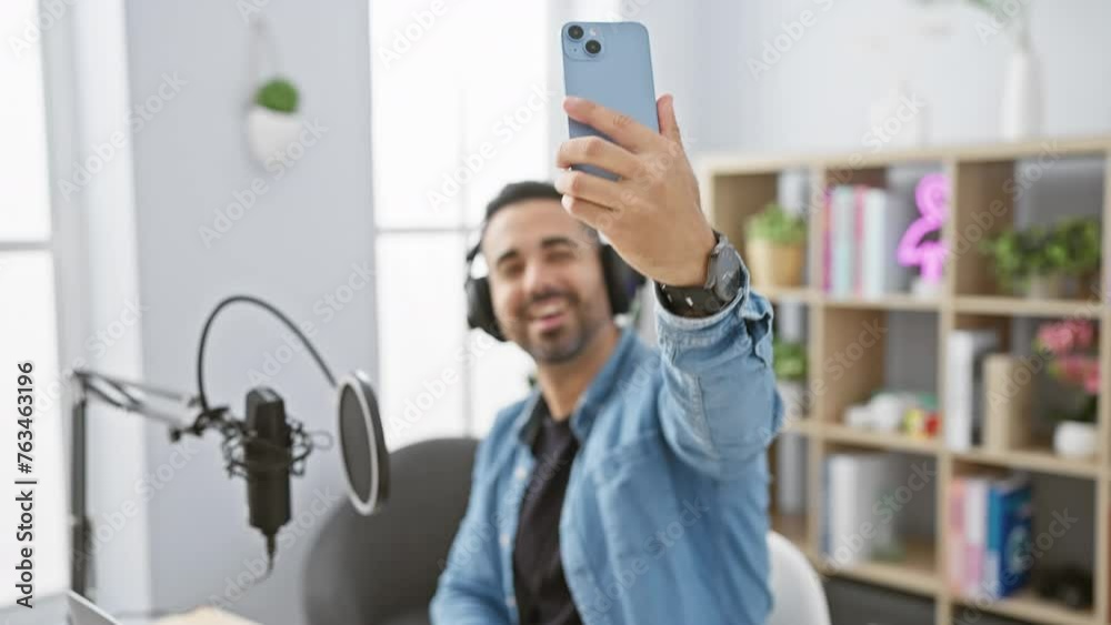 Handsome hispanic man with beard smiling and taking selfie in radio studio using smartphone and wearing headphones. Handsome hispanic man with beard smiling and taking selfie in radio studio using smartphone and wearing headphones.