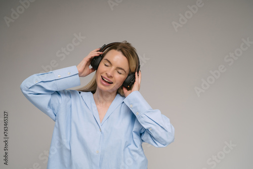 Listening enjoying music. Blonde singing woman with headphones dancing. Studio shot. Gray plain background. Music lover theme banner with copy empty space