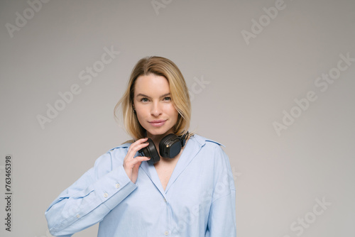 Blonde beautiful young woman with headphones and blue shirt looking at camera. Studio shoot. Gray plain background. Music lover theme banner with copy empty space