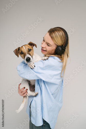 Young beautiful blonde woman music lover with black headphones holding cute dog Jack Russell terrier. Pet looking to the camera. Studio shot, white background