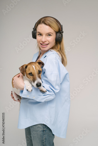 Happy music lover blonde young woman holding small dog Jack Russell terrier looking at camera. Studio shot. Grey background