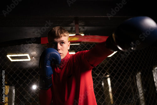 Young handsome boxer in boxing gloves standing near a metal cage in the gym looking at the camera.