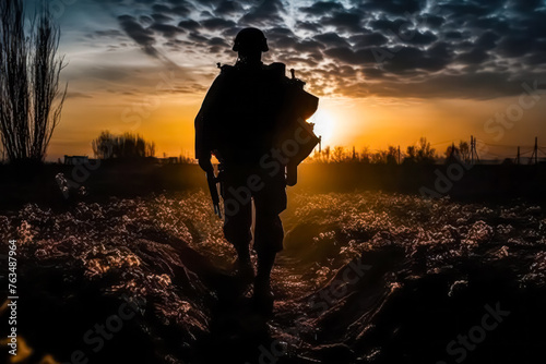 A soldier is walking through a field at sunset. The sky is filled with clouds, and the sun is setting in the distance photo