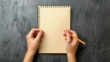 blank light brown notebook with lines, held by two hands and writing in notepad with pencil, gray background, top view.