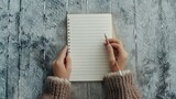 blank light brown notebook with lines, held by two hands and writing in notepad with pencil, gray background, top view.