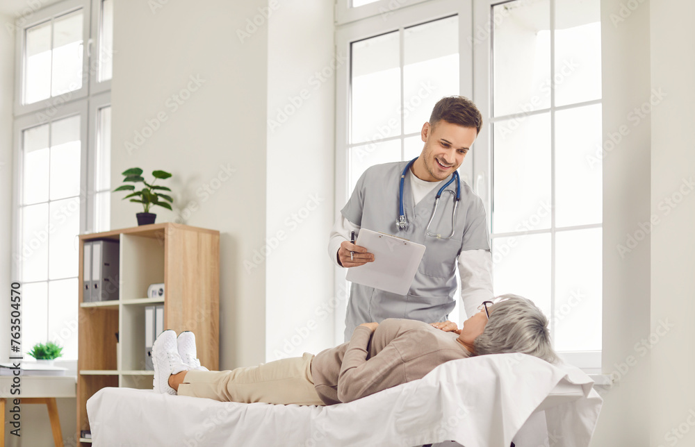 Senior woman patient lies on a couch and talking, consults with a young smiling doctor or nurse in a hospital office. Positive conversation about healthcare in medical clinic environment. Senior woman patient lies on a couch and talking, consults with a young smiling doctor or nurse in a hospital office. Positive conversation about healthcare in medical clinic environment.