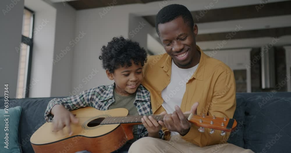 Loving father African American man teaching kid to play the guitar having fun in apartment together. Family and creative activity concept.