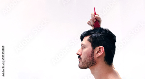 Hombre joven se tiñe el cabello con tinte natural de color castaño oscuro para cubrir sus canas. Tintura a base de henna. Vista de perfil con fondo blanco.  photo