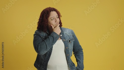 Pensive young woman 20s years old in denin jacket and white t-shirt posing looks around thinks scratches at temple comes up with ideas raised finger up isolated on yellow background in studio. People  photo