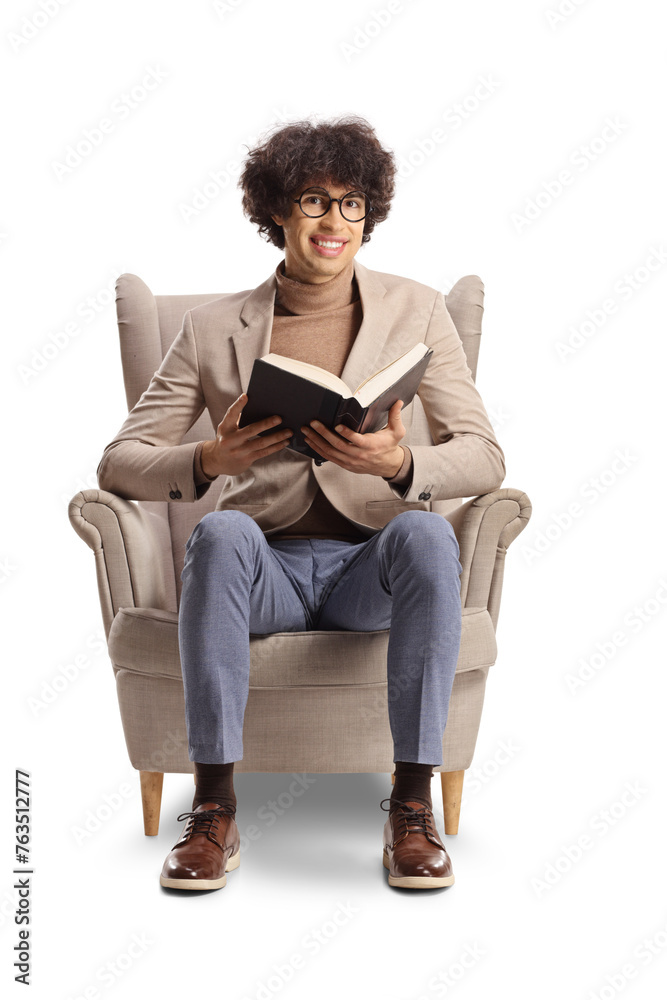 Young man with curly hair and glasses sitting in an armchair and reading a book