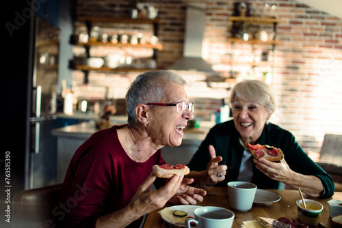 Two senior women laughing and eating breakfast in a home kitchen