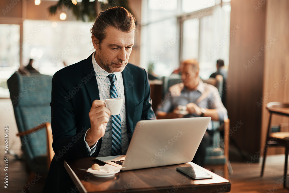 Mature businessman using a laptop at an indoor cafe while having a cup of coffee Mature businessman using a laptop at an indoor cafe while having a cup of coffee