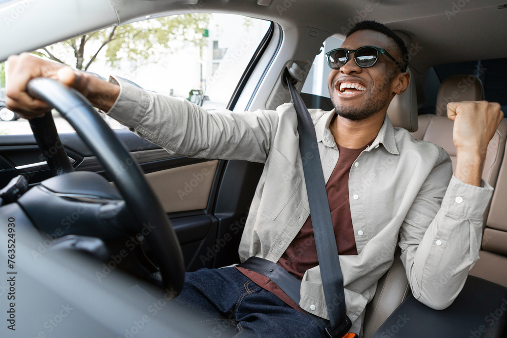Fototapeta premium A joyful african american in glasses dances in a car, sings while driving his car. Road fun