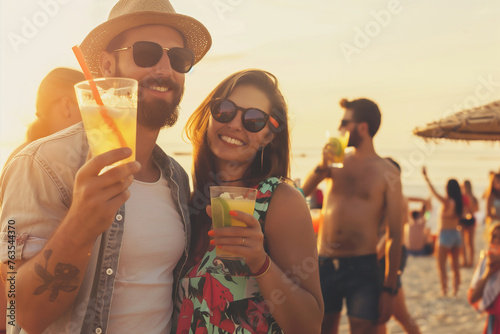 Trendy couple in summer clothing drinking cocktails at beach party. Lifestyle concept with young people having fun at sunset.