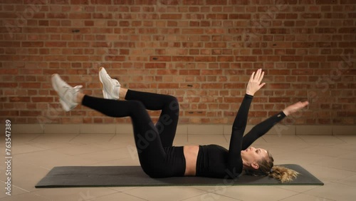 A woman on a gym mat engages her core muscles doing the left-sided dead bug exercise, focusing on controlled movements to enhance stability and abdominal strength in front of a brick wall. Camera RAW.