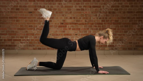 Executing precise donkey kick pulses with her right leg, the professional fitness trainer focuses on glute toning and building endurance, against the structured elegance of a brick-walled gym.
