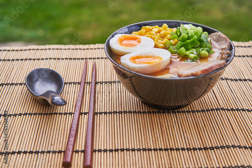 Delicious ramen soup from japan cuisine consist from noodles, pork belly, hard boiled egg, sweet corn seeds and spring onion served in traditional bowl with spoon and chop sticks on bamboo mat outside