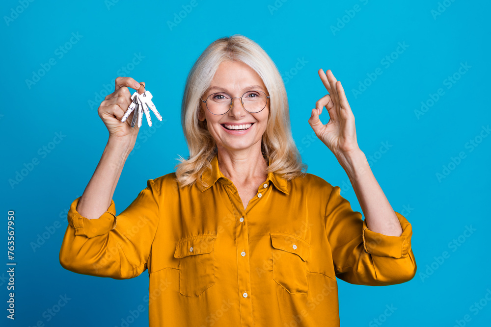 Obraz premium Photo portrait of lovely senior lady hold bunch keys show okey symbol wear trendy yellow silky garment isolated on blue color background