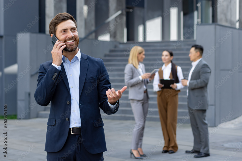 Smiling businessman talking on the phone with a team of professionals engaged in discussion in the background outside a modern office building. Smiling businessman talking on the phone with a team of professionals engaged in discussion in the background outside a modern office building.