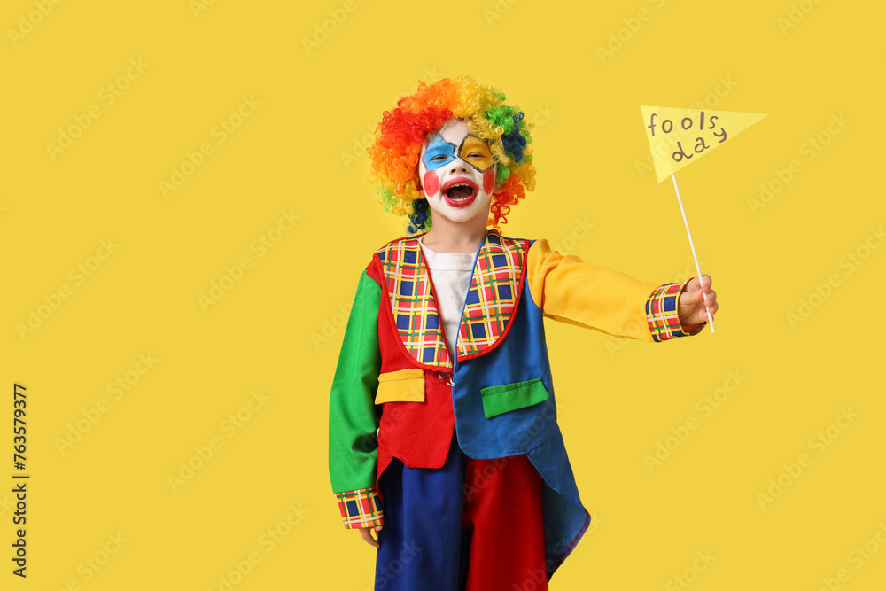Funny little boy in clown costume with paper flag on yellow background. April Fools' Day celebration