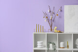 Vase with willow branches, Easter eggs and candles on shelf near lilac wall