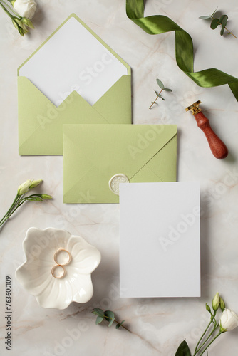 Olive wedding stationery set with blank invitation card, gold rings, wax stamp, flowers on marble table. Flat lay, top view, copy space.