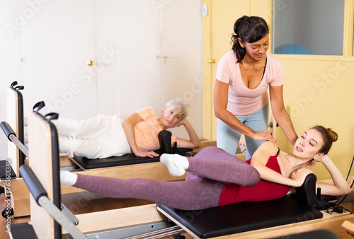 Young woman practicing pilates system on reformer supervised by qualified latin american female trainer..