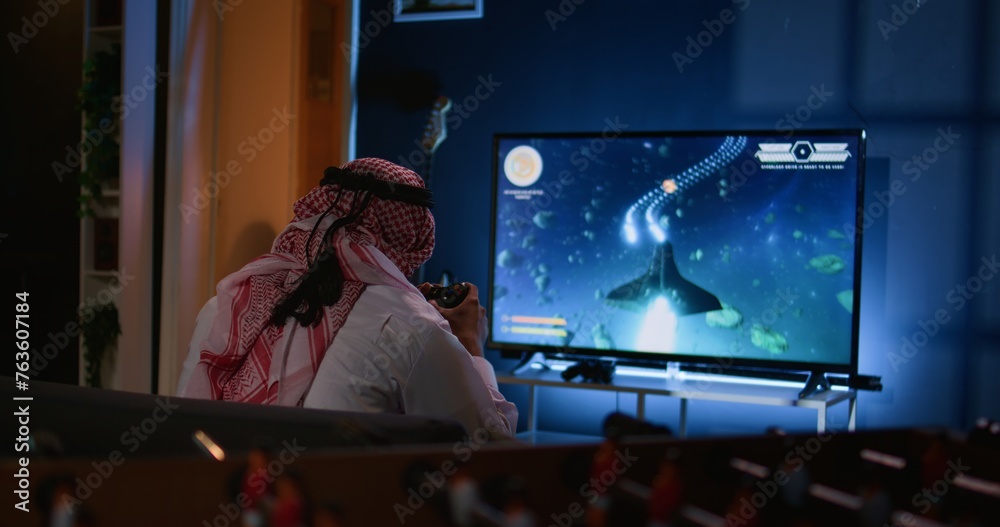 Muslim gamer plays intense classic arcade space shooter videogame, shooting asteroid using laser beams. Arabic man relaxing at home using high tech gaming system to solve missions in singleplayer game