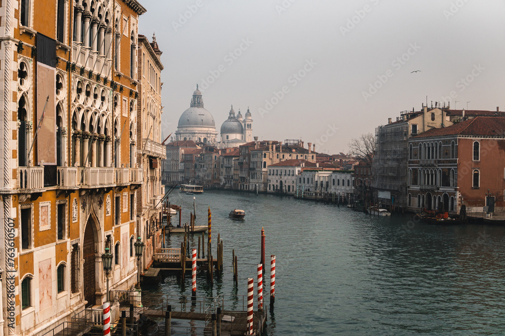Grand Canal from Accademia Bridge: Buildings with medieval-style balconies and windows leading to wooden docks with iconic striped poles. Distant views of Santa Maria della Salute and boats cruising.
