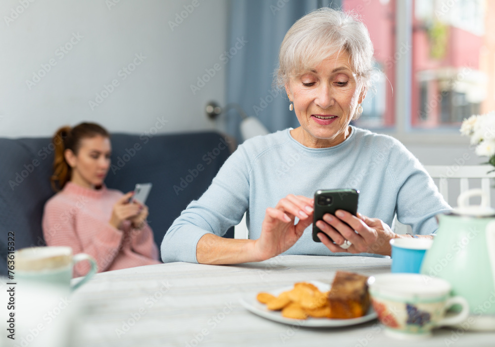 Modern positive elderly woman sitting at table in cozy dining room, using smartphone to chat with friends.