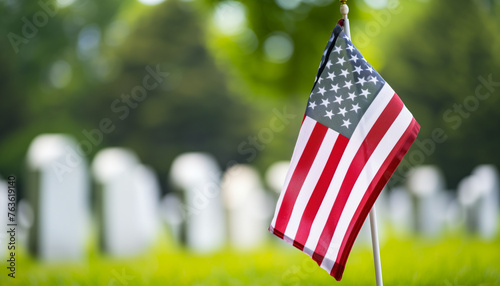 A small American flag is prominently displayed in the foreground with numerous uniform white headstones, indicative of a military cemetery, blurred in the background
