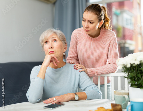 Young girl asking for forgiveness from her offended aged mother sitting at home table with face turned away .
