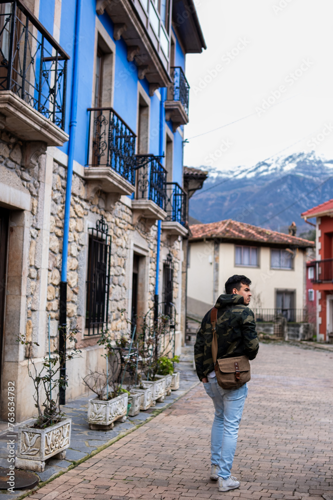 Young Peruvian man in camo coat, brown sling bag, strolls amidst vibrant, stone rural houses.