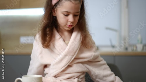 A girl in a bathrobe and a hairband on her head, brought and placed on the table a plate, with two pancake with filling. The child brought breakfast and sat down to eat it