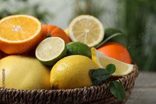 Different fresh citrus fruits and leaves in wicker basket against blurred background, closeup