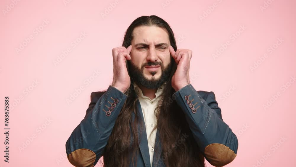 A man with a beard and long hair, wearing a blue jacket and white shirt, grips his temples in distress against a pink backdrop, depicting a headache or mental strain. Camera 8K RAW. A man with a beard and long hair, wearing a blue jacket and white shirt, grips his temples in distress against a pink backdrop, depicting a headache or mental strain. Camera 8K RAW.