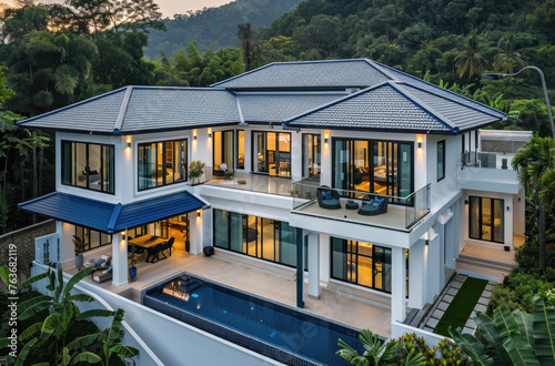 A modern twostory villa with blue tile roof, white walls and glass windows on the first floor. The swimming pool is in front of it, surrounded by lush greenery © Kien