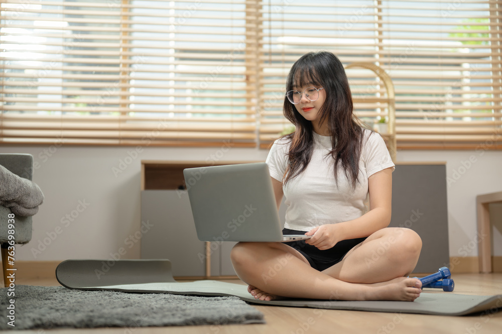 A charming young Asian woman sitting on a yoga mat and using her laptop computer at home.