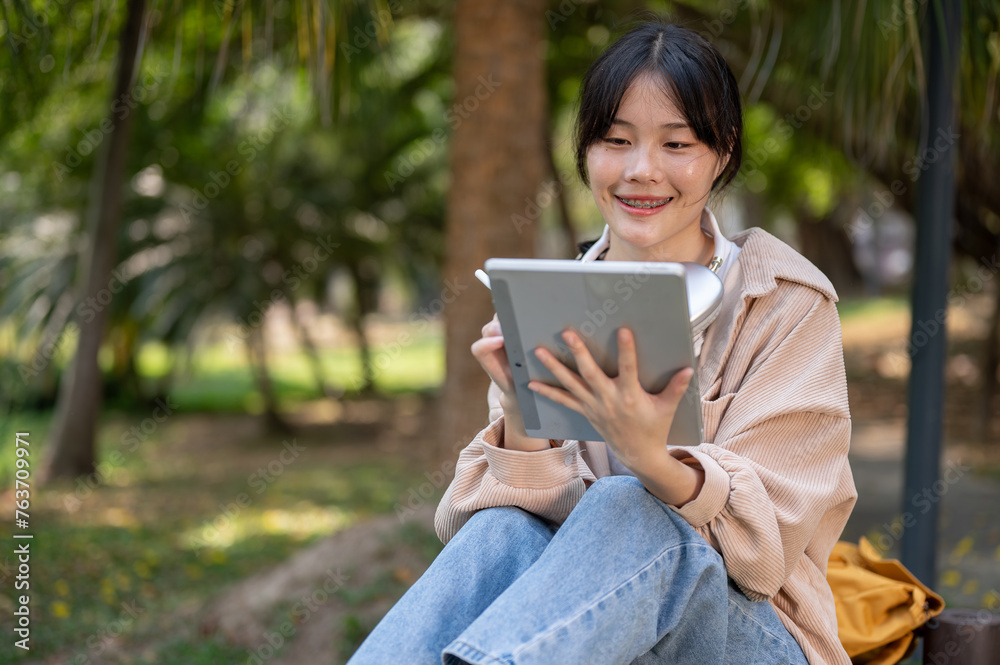A positive, smiling young Asian woman is using her digital tablet on a bench in a park. A positive, smiling young Asian woman is using her digital tablet on a bench in a park.